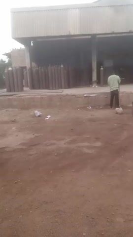 The video shows an industrial yard or facility in daytime, likely in Nigeria. The ground is predominantly dirt or mud. In the foreground, a light-colored scooter or moped is parked on the dirt. A man wearing a light green shirt and dark trousers is standing near a raised concrete platform, facing away from the camera. He is holding a long-handled tool, possibly a shovel or broom, and appears to be moving debris on the ground.

Behind the man and the platform is a large, open-fronted building with a corrugated metal roof. Inside and in front of this building, numerous tall, industrial gas cylinders are neatly arranged. Most of the cylinders are a dark grey or brown color, and some have blue tops, indicating different types of gases. The sky above the building appears overcast. There is some litter on the ground near the platform. The scene suggests a gas filling or distribution plant.