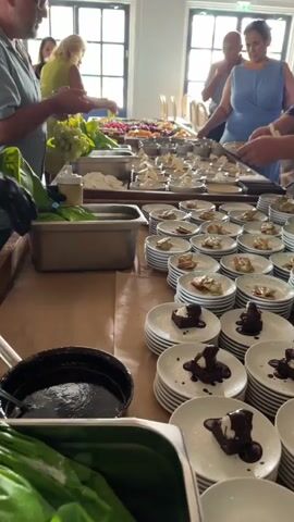 The video shows a buffet line set up indoors, possibly in a catering hall or restaurant. Several people, both male and female, are seen selecting food from a long table laden with various dishes. On the table, there are platters of fruits like grapes, berries, and melon, as well as what appears to be cheeses, bread, and possibly some prepared appetizers. In the foreground, stacks of plates are arranged, some of which already have desserts on them, consisting of small cakes drizzled with sauce and topped with cream. People are actively reaching for food and plates, indicating a meal service in progress. The lighting appears to be natural, coming from windows in the background, with some artificial lighting or colored lights illuminating the dessert plates. The overall atmosphere suggests a catered event or a self-serve meal.