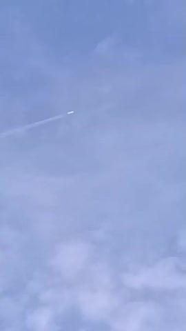 A video shows a distant airplane flying across a light blue sky with scattered white clouds. The plane is leaving a white contrail behind it. The camera is pointed upwards at an angle, capturing the plane as it moves from the left side of the frame towards the upper right. The image is somewhat blurry due to the distance and movement of the plane.
