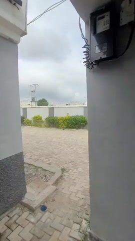 The video shows an outdoor view through a narrow opening between two grey walls. In the foreground, a paved area with interlocking bricks leads to a gravelly section. To the right, an electrical meter box is mounted on a grey wall. Beyond the walls, a low fence with green and yellow shrubs is visible. Further in the background, there are power lines and structures, suggesting an urban or semi-urban environment under a cloudy sky. The scene appears static and without any visible activity.