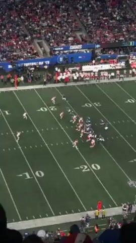 The video shows an American football game from a high, slightly obstructed view in a crowded stadium.  Players in red and white jerseys are on the left side of the frame, while players in blue and white jerseys are on the right, seemingly engaged in a play. Yard lines are visible on the green field. There's a sign that reads "FOOTBALL GIANTS" and part of another sign that says "METLIFE STADIUM." Spectators fill the stands, and a person's arm and the back of their head briefly appear in the lower center of the frame.