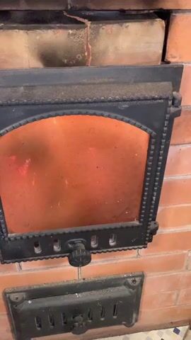 The video opens with a close-up of a closed black metal fireplace door, set into a brick surround. Through the glass window of the door, orange light from glowing embers and fire within is visible. A round knob is centered at the bottom of the door, and another smaller cast-iron grate with a similar knob is situated below it.

The door is then swiftly opened, revealing a blazing fire inside the brick-lined firebox. Large wooden logs are already burning, and new, unburnt logs are introduced into the fire. The flames are bright yellow and orange, actively consuming the wood, and glowing red embers are visible at the base of the fire. The video captures the dynamic movement of the flames as the logs are added.