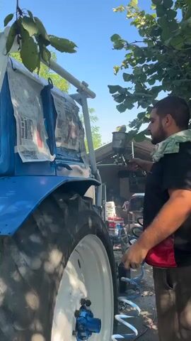 A man with a beard and a towel draped over his shoulders is spray painting a blue tractor. He is holding a spray gun and a blue coiled hose. The tractor's windows are covered with newspaper. The scene is outdoors, with trees and sunlight in the background. The man is wearing a black t-shirt with a red and black ombre design and dark pants. He is focused on his work, carefully applying paint to the tractor's components. The tractor is large and appears to be in the process of being refurbished. The ground is covered in dirt and debris. The overall impression is of a DIY project in progress.