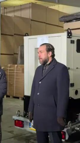 A middle-aged man with a dark beard and mustache, wearing a dark blue overcoat and brown gloves, stands in a warehouse. He is gesturing and speaking, presumably delivering a message. Behind him is a white generator mounted on a trailer, with boxes stacked high in the background.  The environment appears to be a storage or shipping facility.