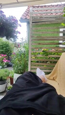A person with white socks on their feet is lying down, covered by a black blanket, in an outdoor patio area. The patio is covered by a white awning and has artificial grass on the floor. There are several potted plants with colorful flowers, including pink and purple petunias, and red roses. A white metal archway stands in the middle of the patio, adorned with a potted rose plant. In the background, there is a two-story brick building with balconies overflowing with flowers. The scene appears to be a private garden or patio, with a relaxed and pleasant atmosphere.