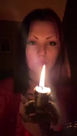 A woman blows out a candle in an indoor environment. She is facing the camera directly, and the lit candle is held at chest level. The light from the candle illuminates her face, which is also lit from a source above and behind her. She is wearing a red top and a necklace. The room appears to be dimly lit, with a picture frame visible on the wall behind her. After blowing, the candle produces a small amount of smoke.