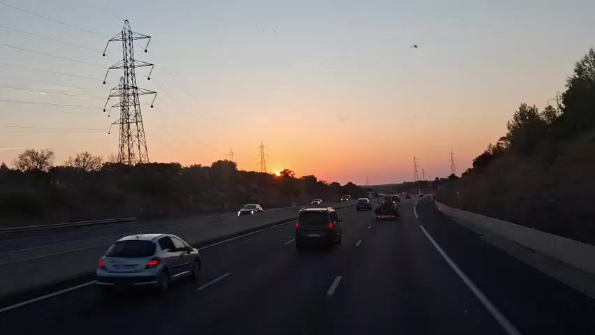 The video shows a highway at sunset. Several cars and trucks are driving on the highway. The sun is setting behind a line of trees on the left side of the road, casting a warm glow over the scene. There are also several large power pylons visible along the side of the highway. The sky is a gradient of orange, pink, and yellow.