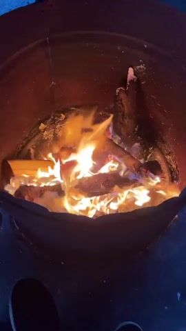 A top-down close-up shot shows a fire burning inside a metal fire pit. Pieces of wood, including logs and smaller branches, are engulfed in bright yellow and orange flames, creating a warm glow. The inside of the fire pit is a mottled red-brown color with some white ash visible. The lower portion of the pit appears dark and undefined, with a few circular holes visible.  The surroundings are generally dark, suggesting the video was taken at night.