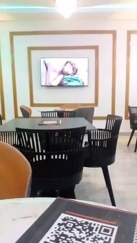 The video shows the interior of a restaurant. The walls are white with brown trim, and there is green lighting along the ceiling. There are multiple tables with a mix of black and orange chairs. Some tables are round and white, while others are rectangular and black. A television is mounted on the wall, and there is an abstract painting on another wall. The floor is tiled.  A menu with a QR code is visible in the foreground.