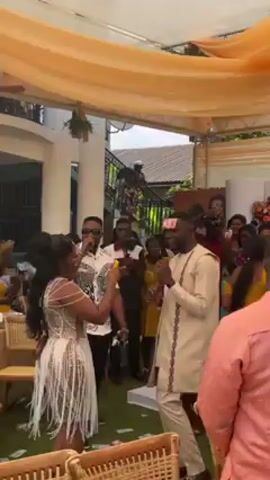 Here is a brief description of the video.

The video shows a wedding reception or similar event.  A bride and groom are dancing in the center of the frame, with a man singing nearby.  The bride is wearing a short, fringed white dress, while the groom is wearing a light-colored traditional outfit.  There are guests seated and standing around, some of whom are recording the event with cameras and phones. Money is scattered on the floor, apparently having been thrown at the couple. The event takes place outdoors under a canopy.