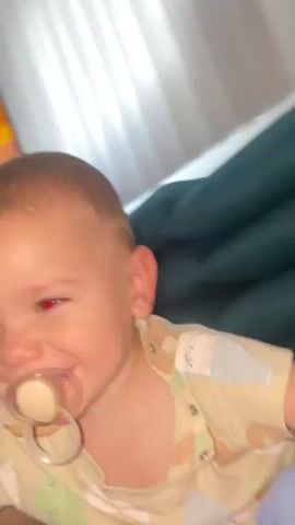 A close-up video shows a baby with a pacifier in its mouth. The baby is wearing a light-colored shirt with cartoon drawings on it. The baby's eyes are brown and it appears to be looking at the camera. In the background, there is a blurred television screen and a curtain. The lighting is dim.