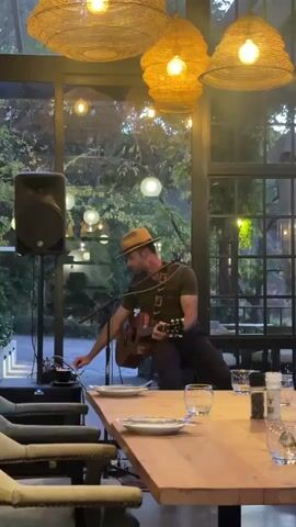 Here's a description of the video:

A man wearing a hat and a dark t-shirt is sitting on a chair on a small stage, playing an acoustic guitar. He adjusts a knob on a small soundboard positioned on the stage. A microphone stands in front of him. A speaker is positioned to his left. He is in a dining room or enclosed outdoor patio with large windows looking out to trees and decorative hanging lights.  A long wooden table with place settings is in the foreground. Woven light fixtures hang from the ceiling.  The space appears empty of other people.