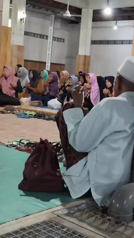 The video shows a group of people, mostly women wearing hijabs, sitting on mats inside a building that appears to be a mosque or community hall. They are facing forward, with their hands raised in a gesture of prayer or supplication. In the foreground, a man wearing a white skullcap and a light blue garment is seated with his back to the camera, also with his hands raised. A maroon bag is visible on the floor next to him. The environment is simple, with bare walls and a ceiling fan. The lighting is artificial, suggesting it is indoors. The overall atmosphere appears to be one of religious observance.