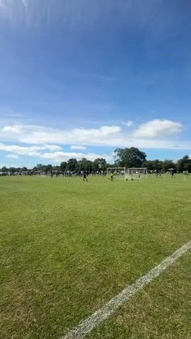 The video shows an outdoor scene on a sunny day with a bright blue sky and scattered white clouds. A large, grassy field is visible, with white boundary lines marking a sports field, likely for soccer. Several people, appearing to be children and adults, are spread across the field, engaged in what seems to be a soccer match or practice. There are at least two soccer goals visible. Some individuals are running, while others appear to be standing or watching. Trees and a line of distant structures are visible in the background. The sun is high in the sky, casting bright light and some lens flare across the footage. The overall impression is of a sporting event taking place in a park or recreational area.
