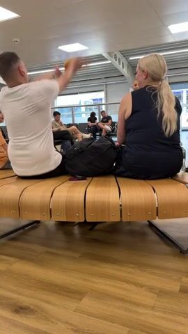 In a medium shot, a man, woman, and child sit on a bench at an airport. The bench is curved and made of light wood slats. The man is wearing a white t-shirt and black shorts, the woman is wearing a black tank top, and the child is wearing a khaki t-shirt. The man and woman are sitting side-by-side, facing forward. The child is standing in front of them, tossing a small stuffed animal in the air. A black backpack sits between the man and woman, and a cell phone is on the bench. Behind them, other people wait in rows of chairs. The airport interior is brightly lit.