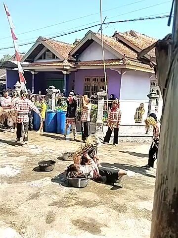 The video depicts an outdoor performance of a cultural dance or ritual. A person wearing a mask and costume is lying on the ground on top of a patterned mat. Several other people stand around, some wearing masks and costumes, others in casual attire. Some of them hold sticks. There are also several small, dark-colored bowls on the ground. In the background, there is a house with a purple exterior and a white porch, as well as blue barrels and what appears to be a red and white striped flag. The ground is concrete and there is a tree partially visible on the right edge of the frame. The scene appears to be taking place during the day under bright sunlight.