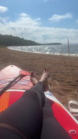 The video shows a close-up shot of a person's legs and feet, resting on a red and white paddleboard. The paddleboard is on a sandy beach, with the ocean and a tree-lined shore in the background. The sky is blue with scattered clouds. The person is wearing black leggings, and their toenails are painted red. A paddle lies next to the person's legs. The sun is shining, and the water is reflecting the light. The scene appears to be a relaxed moment on a sunny day at the beach.