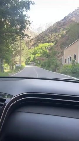 The video shows a car driving on a paved road toward a mountain range. The road is lined with green trees. There is a white car in the distance ahead on the road. There are patches of snow on the mountains in the background. The sky is blue with some clouds. The video is shot from inside the car looking out the windshield. There is a voice heard speaking in a language other than English.