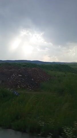 The video shows a landscape scene with a dirt pile in the foreground and rolling hills covered in green vegetation in the background. The sky is overcast with dramatic clouds, and rays of sunlight are breaking through, suggesting either sunrise or sunset. The overall mood is somber or contemplative, enhanced by the muted colors and the raw, undeveloped nature of the terrain. The presence of the dirt pile, possibly from construction or excavation, hints at human activity in the area, juxtaposed with the natural landscape. The time of day appears to be late afternoon or early evening due to the light conditions. There is no visible text in the video. The setting is likely rural, given the open fields and hills.