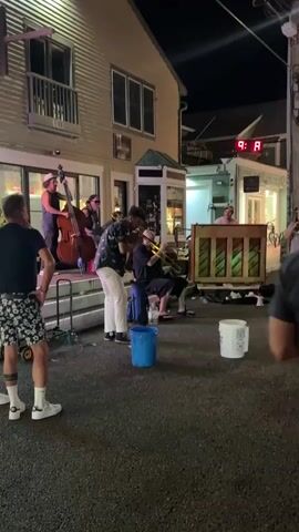 The video shows a street performance by a band at night. In the foreground, a man wearing a black t-shirt and patterned shorts stands facing away from the camera, watching the band. Behind him, on a raised platform, are several musicians. A man in a hat plays a double bass. Another person, possibly a woman, also appears to be on the platform. In front of the platform, a man in a patterned shirt and white pants plays a trombone. Another person, seated, plays a brass instrument, possibly a trumpet or saxophone. A man is seated behind a wooden upright piano on the right side of the scene. The street appears to be paved, and there are buildings on either side. Two buckets, one blue and one white, are visible on the ground in front of the musicians. The lighting suggests it is evening or night, with artificial lights illuminating the scene. There are a few other indistinct figures visible in the background. The overall atmosphere is that of a casual outdoor concert.