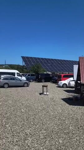 The video shows a wide shot of a parking lot with a gravel surface under a clear blue sky. In the background, a large solar panel array is set up on a sloped structure. Several vehicles are parked in the lot, including a white van, a grey station wagon, a black SUV, a red minivan, and a white sedan. In the center of the shot, a small white table with what appears to be two small objects on it is placed on the gravel. To the right, a wooden structure with a red roof is visible. The overall scene suggests an outdoor area, possibly a business or a residential property utilizing solar energy.