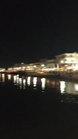 The video shows a nighttime scene of a coastal town. Buildings with illuminated windows line the beach, reflecting on the water. There are a few people visible walking along a paved walkway. The lighting suggests it is evening or night. The camera is moving, giving a sense of being present in the environment.