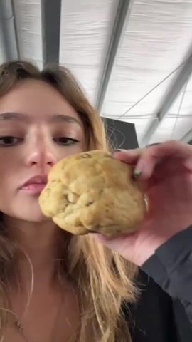 A young woman with long, blonde hair and light brown eyes is holding a large, round cookie to her mouth and taking a bite. She is wearing a black shirt and her nails are painted green. The background is blurry and appears to be an indoor setting with a white ceiling. The lighting is bright and natural.