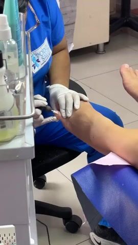 In a medium shot taken indoors, a pedicurist in blue scrubs and white gloves is giving a pedicure.  The scrub top has “PODO Salud Centro Podologico” written on it. The pedicurist uses a small tool to work on the big toe of a client’s foot. The client is sitting on a blue chair with a pink paper barrier. To the left of the frame is a white tiered stand containing bottles and a plastic container of yellow liquid. White tiles cover the floor.  A glimpse of the pedicurist’s black and white Nike shoe can be seen behind the chair.