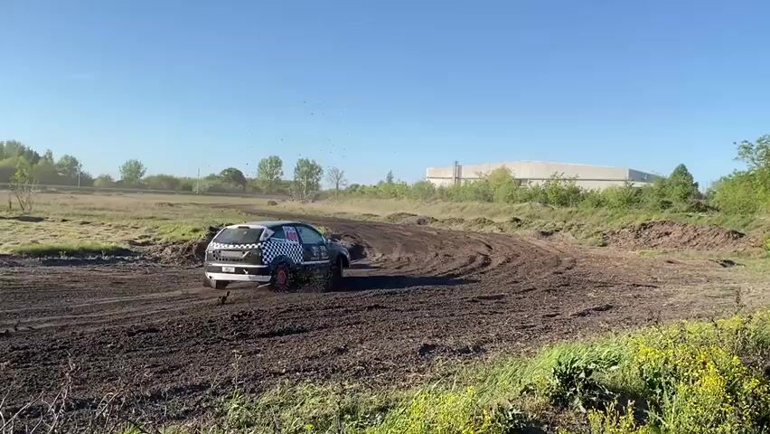 The video shows two cars racing on a dirt track. The first car is a black and white checkered car, and the second car is white. Both cars are kicking up a lot of dust as they navigate the track. The track is located in a field with some trees and a large building in the background. The sky is blue and clear.