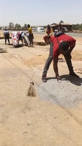 The video shows a group of people working on a dusty road in an outdoor setting.

In the initial scene, two men are working on something on the ground covered in a grey powder. One is wearing a red shirt and jeans, the other is wearing a vest, a cap, and jeans. There is a small box or container near them, as well as a broom to the side. Additional people are seen sitting or standing nearby, observing the activity. In the background, there are buildings, vehicles, and what appears to be a distant hill.

The video then transitions to show more of the street or road. People are visible in the distance sweeping or cleaning the area. They are wearing various outfits. There is a covered structure in the background. 

The video shows a dusty, outdoor environment with people performing cleaning or construction-related tasks.