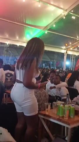 A woman in a white top and shorts dances at a crowded outdoor event under a large tent with string lights. She faces away from the camera, moving near a wooden table covered with plastic cups and cans. Other people are seated or standing around tables in the background. In the distance, a stage structure is visible. The lighting changes color throughout the short clip.