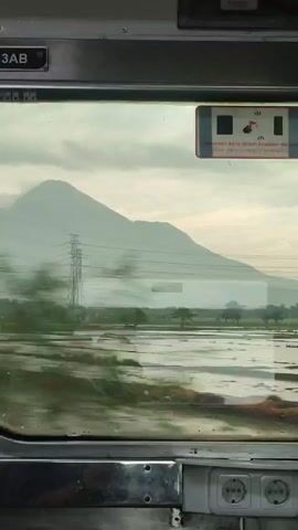 Here is a description of the video:

The video shows a view from a train window. In the distance, a large mountain looms under a slightly overcast sky. Two tall electrical towers stand between the train and the mountain, with power lines stretching across the landscape. The immediate foreground outside the window consists of flooded fields, likely rice paddies, with some green vegetation visible within the water.  The train is moving, which is evident from the blurring of the foreground landscape. Inside the train compartment, a seat number "3AB" is visible above the window. A small sticker with instructions (in Indonesian) about breaking the glass in case of emergency is located on the right side of the window.  Below the window, there are two electrical outlets, indicating that this is the interior of a passenger train.