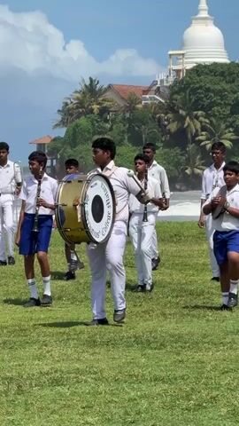A group of young males in white school uniforms with blue shorts are marching in formation on a grassy field. They are carrying musical instruments, including clarinets and a bass drum. The person in the foreground is playing the bass drum, while others are holding clarinets and a trumpet. In the background, there is a body of water, possibly the ocean, with waves crashing on the shore. Further in the distance, there is a white dome-shaped building, possibly a Buddhist temple, situated on a hill with palm trees. The sky is blue with scattered white clouds. The scene appears to be a musical performance or parade in an outdoor setting.