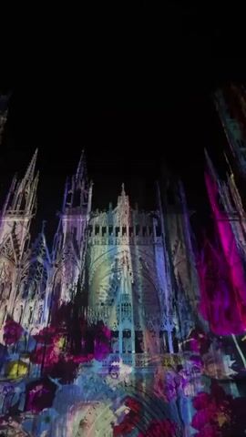 The video depicts a tall, ornate gothic cathedral at night. The cathedral is illuminated by an animated projection mapping display that casts a variety of vibrant colors and patterns onto its facade. The colors change and move, creating a dynamic light show. The patterns include vertical stripes of shifting hues and abstract painterly strokes. The video is shot from a low angle, looking up at the cathedral against a dark night sky.