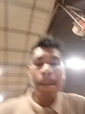 A man is recording a selfie video in an indoor space. The man has short, dark hair styled in a mohawk. He is wearing a tan collared shirt. The room has a ceiling made of white panels with dark metal frames, and there are several round lights overhead. The man looks directly at the camera, his face relatively still. The camera is hand-held and moves slightly.