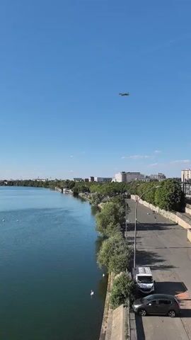 The video shows a view from a bridge overlooking a wide river under a clear blue sky. An airplane is visible in the distance, flying high above. On the right side of the river, there is a road with a few parked cars and some trees lining the bank. Further down the road, there are buildings and more trees. On the left side of the river, there are more trees and some buildings along the opposite bank. Several white swans are seen swimming in the river. The overall scene appears to be a peaceful day in an urban or suburban area with a prominent waterway.