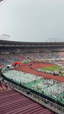 The video shows a large stadium filled with people, viewed from an elevated angle. The stadium is packed with spectators in the stands, and the field is filled with rows of white chairs, many of which are occupied. A running track surrounds the field. There are structures set up on the field, including a stage and large screens displaying images. A significant number of people on the field are wearing white tops, possibly as part of a uniform or event theme. The sky is overcast, creating a gray and diffused light.