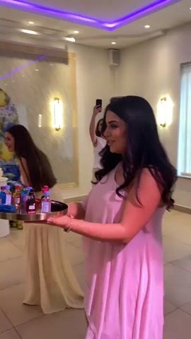 In a well-lit, decorated interior, three young women participate in an event. One woman in a pink dress holds a round tray with miniature liquor bottles, walking toward others who are reaching to take a bottle. Behind her, another woman in a light yellow dress laughs, while the third, in a dark brown dress, takes a bottle from the tray. A fourth woman in a white top and tan pants stands farther back, holding a phone up as if taking a video or photo.

Behind the group, there is a decorative screen with a blue and white pattern and yellow fruit, a white multi-tiered cake on a pedestal, two sconces on the wall, and a large speaker on a stand next to a laptop. Overhead, blue and green strip lights provide additional lighting to the event space. The floor is tiled.