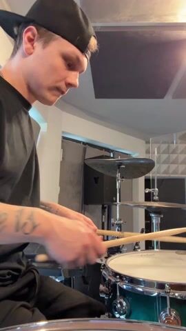A male drummer is playing a drum kit in a recording studio. The man is fair-skinned with short blonde hair and a black baseball cap worn backwards. He is wearing a black t-shirt, and visible on his forearm are tattoos. He is smiling and making eye contact with the camera as he plays. The drum kit is partially visible, with a dark green snare drum in the foreground. Behind the snare drum are cymbals. The studio has soundproofing panels on the walls and ceiling. There is a door in the background.