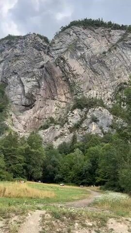 The video pans from left to right, showcasing a large, rocky mountain face with a thin waterfall cascading down from the top. In the foreground, there is a lush green grassy field with scattered wildflowers. Several trees with dense green foliage are present, bordering the field and the mountain. A dirt path runs through the grassy area, and a few people are seen walking along it. The sky is overcast with dramatic clouds, suggesting potentially changing weather. The scene is set in a natural, mountainous environment.