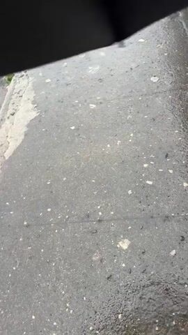 The video begins with a blurred shot of a wet surface, likely pavement or asphalt, with scattered debris. It then pans to reveal a weathered concrete structure, possibly a bridge or walkway, with moss or algae growing on it. The camera then moves to capture a wide view of a fast-flowing river. The water is muddy brown, indicating turbulence and a high volume. Trees and green foliage are visible on the riverbanks, suggesting a natural or semi-natural environment. The water is choppy with visible waves and currents, indicating a powerful flow. The overall scene depicts a flood event or a river experiencing very high water levels, likely due to heavy rainfall. There are no people visible in the video.