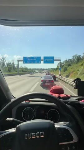 The video is taken from the driver's perspective inside a vehicle, looking forward through the windshield. The vehicle is on a multi-lane highway with a clear blue sky and some sun glare. There are other vehicles on the road, indicating traffic. Overhead signs direct traffic to Karlsruhe, Basel, Pforzheim-Süd, Heimsheim, and Mönsheim. A red, plush item is visible on the dashboard. The steering wheel of the vehicle, with the IVECO logo, and part of the dashboard with a speedometer and tachometer are in the foreground. Trees and vegetation line the side of the highway. The overall scene depicts driving on a highway with moderate traffic.