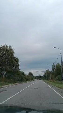A car is driving down a paved road under an overcast sky. The road has two lanes with a white line marking the center. There are trees and bushes on either side of the road. The view is from inside the car, showing the dashboard and the windshield. The camera pans slightly to the right at one point, showing the side mirror.