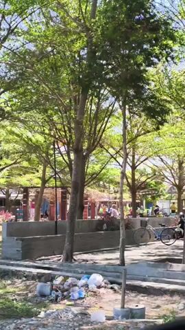 The video shows an outdoor scene with several trees providing shade. In the foreground, there is a pile of garbage. Behind the garbage and trees, there is an area with people and stalls, suggesting a market or outdoor gathering. Two bicycles are visible, one of which is being ridden by a person. There are also other people walking around in the background. The overall environment appears to be sunny.