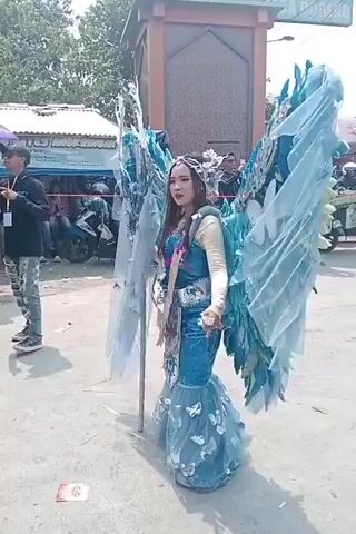 A young woman wearing a large, ornate costume with blue, feathered wings walks down a dirt path. She has on a long, blue, mermaid-style dress with a ruffled hem adorned with butterflies. Her top is a light-colored long-sleeved shirt, and she has a sash draped across her chest with what appears to be money pinned to it. Her hair is dark and appears to be styled with a headband or crown. Behind her, there are several stalls or tents set up, with people milling about. A motorcycle is parked to the left. To the right, a group of people are standing, some appearing to be taking photos. A tall, ornate structure with a peaked roof is visible in the background. The overall environment appears to be an outdoor market or festival on a sunny day.