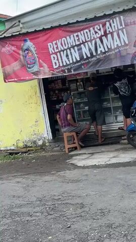 The video shows the exterior of a small shop. A red banner with white text reads "REKOMENDASI AHLI BIKIN NYAMAN" which translates to "Expert recommendation makes it comfortable". Below this text in smaller font, it says "Elektro kurang Gesekan Mesin Matic, Tarikan Enteng Mantap" meaning "Electric motor less friction, Smooth acceleration, Great". A man sits on a stool to the left of the shop's entrance. Two men are inside the shop, one appearing to be working on something near the back. A blue and black motorcycle is parked in front of the shop to the right. The shop has shelves stocked with various items visible through a glass window. The ground in front of the shop is paved and appears somewhat worn.