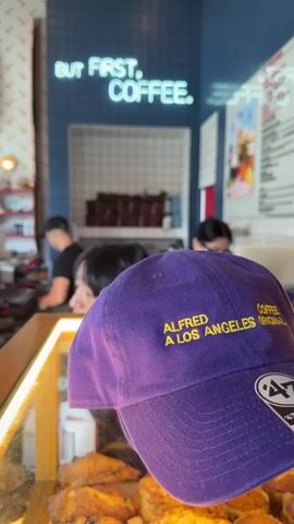 The video opens with a close-up of a purple baseball cap with gold lettering that reads "ALFRED A LOS ANGELES COFFEE ORIGINAL". The camera then pans to the left, revealing the interior of a brightly lit coffee shop. A neon sign on the back wall displays the words "BUT FIRST, COFFEE." In the foreground, behind a glass display case filled with pastries, two individuals are visible. Further back, a barista wearing a black t-shirt and apron is preparing drinks behind a counter. Another person, likely a customer, is standing at the counter. The shop's decor includes blue tiled walls, shelves with coffee bags and merchandise, and white checkered tiling. A man with dark hair is working at a coffee machine. The overall atmosphere appears to be busy and casual.