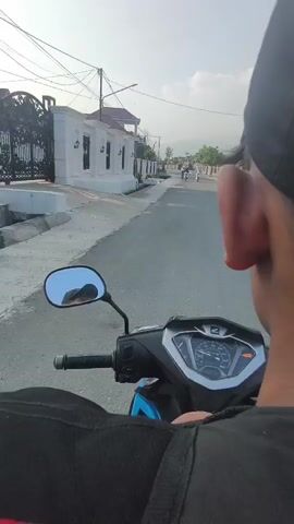The video shows a first-person perspective of someone riding a motorcycle down a paved road. The rider is wearing a black cap and a black backpack. In the foreground, the motorcycle's handlebars and dashboard are visible. The road is lined with houses and trees on both sides. In the distance, a group of people are riding bicycles, also moving down the road. The sky is clear with some clouds, suggesting it is daytime.