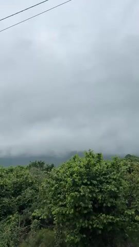 The video depicts a cloudy day in a rural or semi-rural area. The foreground features a rusty red corrugated metal fence. Behind the fence are green trees and shrubs. In the distance, partially obscured by the low-hanging clouds or fog, are hills or mountains. A house with a red roof is visible to the left of the frame. There are also electrical wires in the top of the frame. The camera pans upward, focusing more on the overcast sky and the red fence.