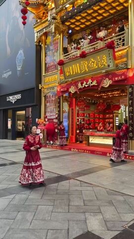 A video shows the facade of a jewelry store, "Sichuan Gold Store," decorated in a traditional Chinese style with gold accents and red banners. Women in red traditional dresses stand in front, with some holding red decorations. There's a crowd of people visible on a balcony area. To the side, there's a Longines watch advertisement on a modern building. The general scene is vibrant and festive.